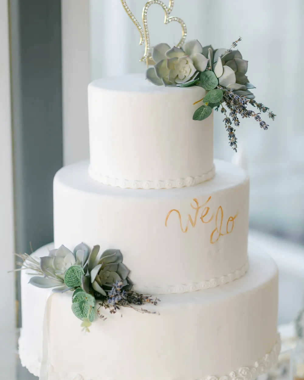 A three-tier white wedding cake with succulents and gold script