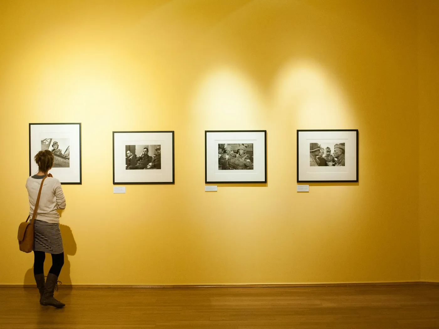 Framed works hung along a warm yellow gallery wall, a visitor looking on
